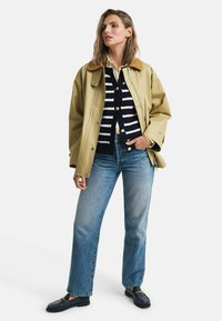 Kaki vattentålig jacka med brunt krage, marinrandig knappad cardigan, ljusblå raka jeans och mörka loafers med kedjeaccent.