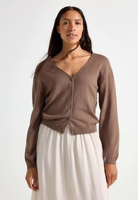 Brown knit cardigan with a V-neck, long sleeves, and front buttons. Soft texture with a fitted waistband, worn over a white skirt.