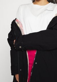 Svart ytterjacka med en vadderad rosa insida, lager över en vit t-shirt. Har dragkedja och tryckknapplås.