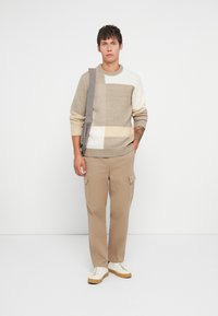 Ανδρικό πλεκτό πουλόβερ σε μπεζ με σχέδιο σε χρωματική μπλοκ, με ανοιχτές και σκούρες περιοχές. Συνδυασμένο με μπεζ cargo παντελόνια και λευκά αθλητικά παπούτσια.