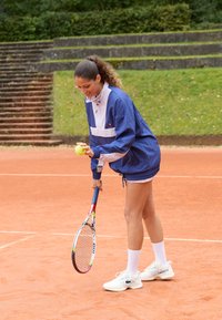 Frau in blauer Jacke hält einen Tennisball und einen Schläger und bereitet sich darauf vor, auf einem Tennisplatz aus roter Erde zu servieren, mit Grünflächen und Stufen im Hintergrund.