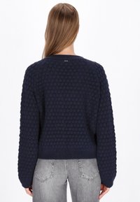 Granatowy sweter z teksturowanym, bąbelkowym splotem, ściągaczem u dołu i długimi rękawami. Materiał wygląda na miękki i przytulny. Pokazany widok z tyłu.