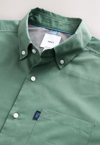 Camicia verde con bottoni in cotone liscio, con taschino sul petto e bottoni bianchi. Presenta un dettaglio blu all'interno del colletto. Vestibilità regolare.