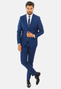 Completo blu con pantaloni abbinati, camicia bianca e cravatta scura. Presenta un design elegante e una silhouette aderente, abbinato a scarpe nere lucide.