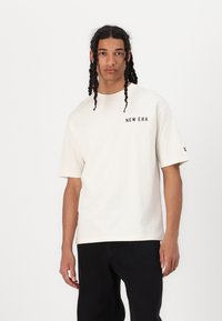 Kortærmet, cremegul bomulds T-shirt med "NEW ERA" sort logo på brystet. Løs pasform og rund halsudskæring. Sættet er parret med sorte bukser.