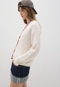 Hellrosa gerippte Strickjacke aus weichem Material, getragen über einem gestreiften Oberteil, kombiniert mit dunkelblauen Denimshorts mit umgeschlagenem Saum und Akzentstreifen.