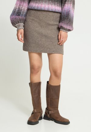 Jambes portant des bottes hautes marron, une jupe en laine marron et un pull en maille multicolore à manches bouffantes sur un fond neutre.