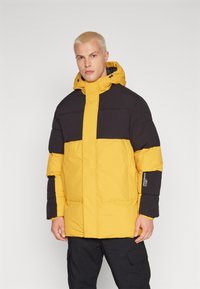 Chaqueta acolchada con una sección superior negra y una inferior amarilla. Presenta una capucha, cierre de cremallera y puños elásticos. Tejido suave y texturizado.