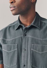 Camicia a maniche corte di colore grigio scuro con tessuto strutturato, dotata di due tasche sul petto, bottoni neri e cuciture bianche a contrasto.