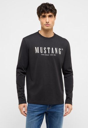 Mann trägt ein schwarzes Langarmshirt mit dem Schriftzug "MUSTANG Jeans Company Since 1932", dazu blaue Jeans, steht vor einem weißen Hintergrund.