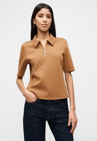 Γυναίκα που φοράει πόλο πουκάμισο κοντομάνικο σε χρώμα καμήλας με φερμουάρ μπροστά και σκούρο μπλε τζιν, όρθια με το ένα χέρι στην τσέπη.