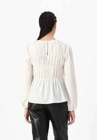 Blusa bianca a maniche lunghe con schiena arricciata, scollatura tonda e dettaglio a occhiello. Il tessuto leggero crea una silhouette morbida e fluida.