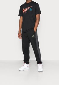 Musta Nike t-paita, jossa on valuvan logon muotoilu, yhdistettynä mustiin collegehousuihin, joissa on vihreä "AIR"-korostus. Valkoiset tennarit viimeistelevät asun.