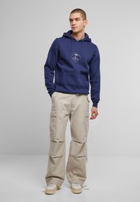 Felpa con cappuccio blu navy dotata di tasca frontale e logo; abbinata a pantaloni cargo beige chiaro con tasche laterali e orli regolabili.