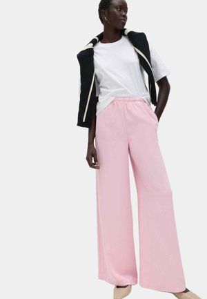 Kobieta w białej koszuli z krótkim rękawem, czarnym swetrze zarzuconym na ramiona, różowych spodniach z szerokimi nogawkami i beżowych, szpiczastych butach, stojąca z ręką w kieszeni.