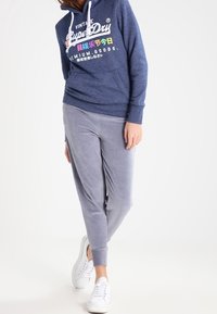 Felpa navy con cappuccio e lacci bianchi, abbinate a pantaloni jogger grigi chiari, entrambi in tessuti morbidi. Sneakers bianche completano il look.