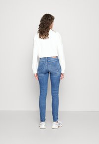 Jeans ajustados de mezclilla en azul, con bolsillos traseros de parche y una cintura alta, combinados con un suéter blanco texturizado y zapatillas blancas.