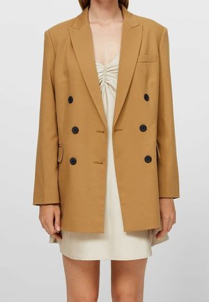 Vrouw met een dubbelgeknoopte blazer in kamelkleur over een crèmekleurige mini-jurk met ruches, staand tegen een effen lichte achtergrond.