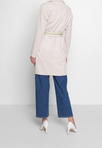 Beige mockaskä coat med bältesmidja, långa ärmar och krage, kombinerad med vida blå denimbyxor och vita klackar.