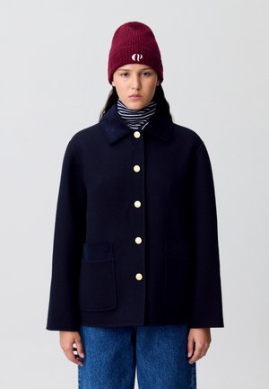 Manteau en laine marine avec une fermeture à boutons jaunes, deux poches avant et un col en velours côtelé, associé à un col roulé rayé et un bonnet bordeaux.