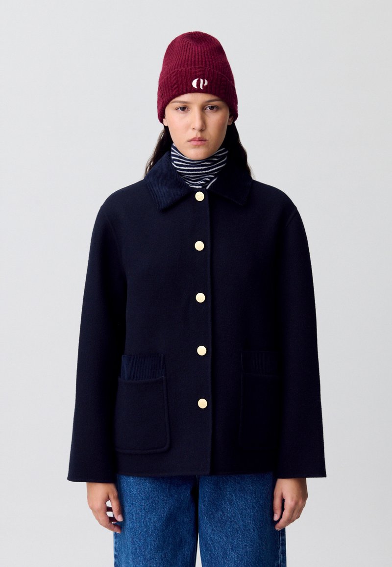 Cappotto di lana navy con chiusura a bottoni gialli, due tasche frontali e colletto in velluto a coste, abbinato a un dolcevita a righe e a un berretto bordeaux.