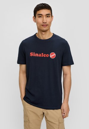 Dunkelblaue Baumwoll-T-Shirt mit einem roten "Sinalco"-Logo und runder Akzent. Klassisches Rundhalsdesign. Getragen mit khakifarbenen Cargo-Hosen.