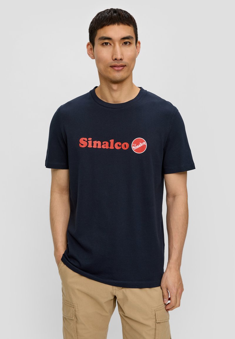 Tamsiai mėlynas medvilninis marškinėlis su raudonu "Sinalco" logotipu ir apvaliomis detalėmis. Klasikinio apvalaus kaklo dizainas. Dėvimas su chaki spalvos krovinių kelnėmis.