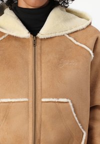 Veste en suède beige avec une capuche doublée de shearling, dotée d'une fermeture éclair et de deux poches avant. Détail de logo discret sur la poitrine.