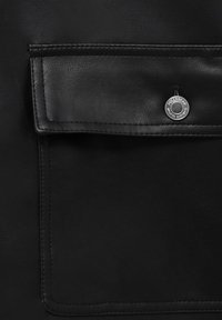 Gros plan sur une poche en cuir noir avec un rabat fixé par un bouton rond en argent gravé des mots "Pull&Bear" et "Unlimited Garments".