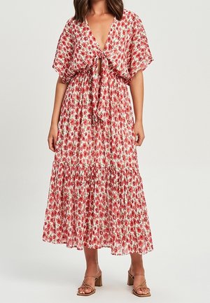 Femme portant une robe midi fleurie à manches courtes avec un noeud devant, associée à des sandales marron à brides et talons épais.