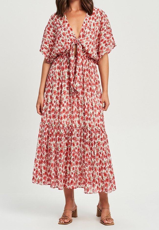 Femme portant une robe midi fleurie à manches courtes avec un noeud devant, associée à des sandales marron à brides et talons épais.