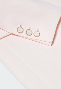 Poignet de chemise rose clair avec trois boutons de manchette ronds blancs et dorés attachés en ligne.