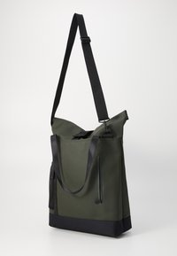 Olivgrüne Tote-Tasche mit strukturiertem mattem Finish, ausgestattet mit einem oberen Verschluss, verstellbarem schwarzen Trageriemen und einer seitlichen Netztasche.