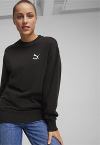 Felpa nera con colletto rotondo, con il logo bianco Puma sul petto, abbinata a jeans blu. Tessuto morbido e vestibilità rilassata.