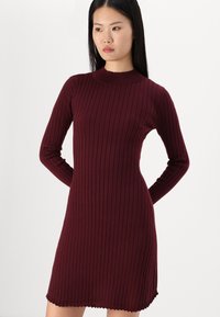 Vestido bordeaux com design ajustado, mangas longas, gola alta e bainha em formato de ondas. Feito de um material de malha texturizada.