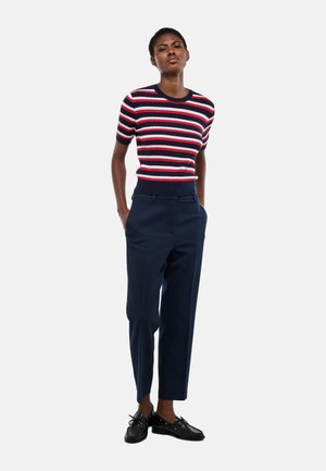 Femme debout, les mains dans les poches, portant un pantalon bleu marine, des mocassins noirs et un haut en tricot à manches courtes rayé rouge, blanc et bleu, sur fond blanc.