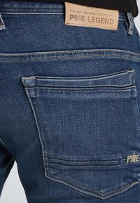 Traperice u tamnoplavoj boji, sa kožnom oznakom označenom "PME LEGEND", kontrastnim šavovima i dizajnom stražnjeg džepa.