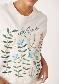 Sweatshirt beige clair avec des motifs floraux brodés en vert, bleu, orange et jaune. Col ras du cou et manches courtes.