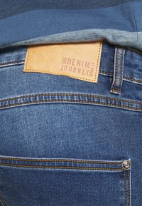 Jeans in denim con un'etichetta in pelle contrassegnata "THE DENIM JOURNEY" e cuciture arancioni; toni blu e texture visibili; dettaglio con bottone sulla tasca.