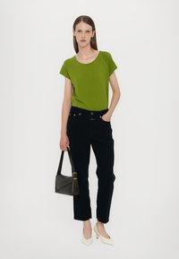 Green short-sleeve top paired with dark jeans; model holds a dark handbag. White pointed-toe shoes complete the outfit. Plain background.
