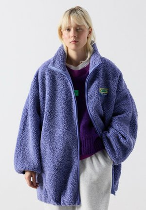 Junge Person trägt eine übergroße, strukturierte violette Fleecejacke, violetten Pullover und hellgraue Hose vor einfachem Hintergrund.