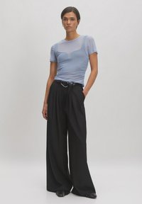 Lichtblauw fitted top met korte mouwen; doorschijnende textuur. Zwarte wijde broek met een riem. Model staat tegen een neutrale achtergrond.