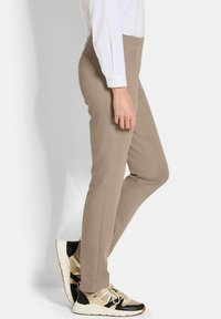Persoon in beige slim-fit broek, wit overhemd met lange mouwen, en beige en zwarte sneakers, staand tegen een effen witte achtergrond.