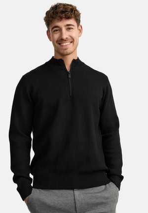 Suéter negro de punto con media cremallera, que presenta un cuello y puños acanalados. El material parece suave; se lleva sobre pantalones claros de cuadros grises.