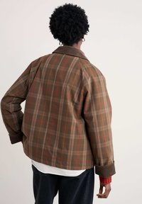 Brūns raibs žakets ar korduroja apkakli, kas ietver sarkanas un brūnas akcentus. Audumam ir gluda tekstūra un brīva piegriezuma.