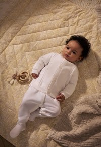 Cardigan blanc en tricot avec un motif texturé, associé à un pantalon blanc. Bébé allongé sur une couverture jaune à motif floral avec un anneau de dentition en bois à proximité.