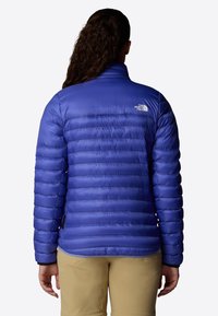 Giacca isolante blu con design trapuntato orizzontale, colletto alto e logo sulla parte superiore della schiena. Realizzata in materiale lucido e leggero.
