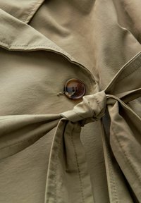 Tessuto beige con una superficie liscia, caratterizzato da un grande bottone marrone e un dettaglio di cintura annodata in vita.