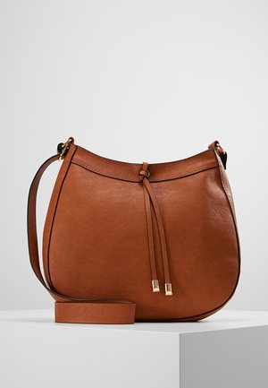Sac bandoulière en cuir marron avec une sangle réglable et un nœud décoratif devant, présenté sur un socle blanc sur fond uni.