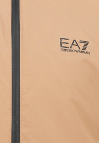 Šviesiai smėlinė striukė su lygiu paviršiumi, turinti juodą užtrauktuką ir juodą EA7 Emporio Armani logotipą kairėje pusėje.